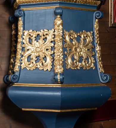 Zdjęcie nr 3: Ambona przyścienna, wisząca z dojściem od zakrystii. Korpus wieloboczny, rozszerzający się ku dołowi, z narożnikami zaakcentowanymi długimi spływami z dekoracją rzeźbiarską, kwiatową, zakończonymi wolutami. Pola korpusu ozdobione ażurowymi, kwadratowymi rozetami; parapet w formie profilowanego, wydatnego gzymsu; dolne zakończenie korpusu w kształcie wybrzuszenia, przechodzącego dołem w ostrosłup ścięty, o wklęsłych bokach, zakończony puklowaną szyszką. Baldachim pięcioboczny, ujęty wysokim, profilowanym gzymsowaniem, z lambrekinem od dołu, zwieńczony ażurowymi esownicami spiętymi u szczytu wazonem płomienistym. Pomiędzy spływami wolutowymi akroteriony. Na podniebiu rzeźbiona gołębica Ducha Świętego na tle promienistej glorii. Struktura malowana na kolor ciemnoniebieski, ornamenty i profile złocone. Drzwi na ambonę nowe.