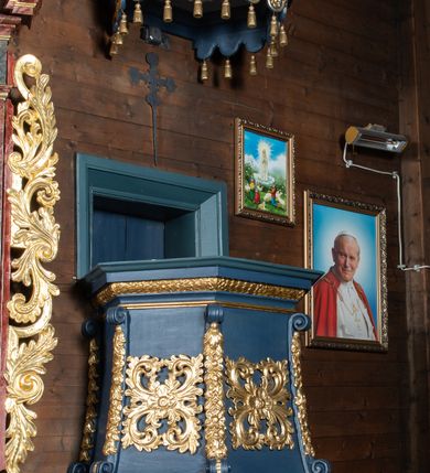 Zdjęcie nr 2: Ambona przyścienna, wisząca z dojściem od zakrystii. Korpus wieloboczny, rozszerzający się ku dołowi, z narożnikami zaakcentowanymi długimi spływami z dekoracją rzeźbiarską, kwiatową, zakończonymi wolutami. Pola korpusu ozdobione ażurowymi, kwadratowymi rozetami; parapet w formie profilowanego, wydatnego gzymsu; dolne zakończenie korpusu w kształcie wybrzuszenia, przechodzącego dołem w ostrosłup ścięty, o wklęsłych bokach, zakończony puklowaną szyszką. Baldachim pięcioboczny, ujęty wysokim, profilowanym gzymsowaniem, z lambrekinem od dołu, zwieńczony ażurowymi esownicami spiętymi u szczytu wazonem płomienistym. Pomiędzy spływami wolutowymi akroteriony. Na podniebiu rzeźbiona gołębica Ducha Świętego na tle promienistej glorii. Struktura malowana na kolor ciemnoniebieski, ornamenty i profile złocone. Drzwi na ambonę nowe.