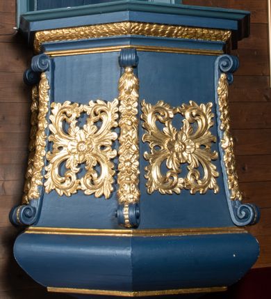 Zdjęcie nr 1: Ambona przyścienna, wisząca z dojściem od zakrystii. Korpus wieloboczny, rozszerzający się ku dołowi, z narożnikami zaakcentowanymi długimi spływami z dekoracją rzeźbiarską, kwiatową, zakończonymi wolutami. Pola korpusu ozdobione ażurowymi, kwadratowymi rozetami; parapet w formie profilowanego, wydatnego gzymsu; dolne zakończenie korpusu w kształcie wybrzuszenia, przechodzącego dołem w ostrosłup ścięty, o wklęsłych bokach, zakończony puklowaną szyszką. Baldachim pięcioboczny, ujęty wysokim, profilowanym gzymsowaniem, z lambrekinem od dołu, zwieńczony ażurowymi esownicami spiętymi u szczytu wazonem płomienistym. Pomiędzy spływami wolutowymi akroteriony. Na podniebiu rzeźbiona gołębica Ducha Świętego na tle promienistej glorii. Struktura malowana na kolor ciemnoniebieski, ornamenty i profile złocone. Drzwi na ambonę nowe.