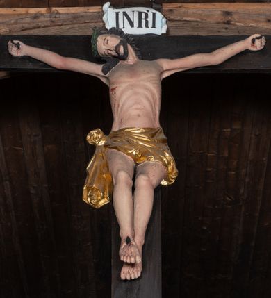 Zdjęcie nr 1: Krucyfiks z rzeźbą Chrystusa w typie Christo morte. Ciało zawieszone na niemal wyprostowanych ramionach; głowa silnie przechylona na prawe ramię; nogi delikatnie ugięte w kolanach, stopy przebite jednym gwoździem w układzie prawa na lewą. Twarz podłużna z zamkniętymi oczami i długim nosem, okolona krótkim zarostem. Na głowie zielona korona cierniowa. Włosy długie, czarne, opadające na prawe ramię oraz na plecy, silnie zawinięte do tyłu w drobne pukle. Klatka piersiowa szeroka z delikatnie, łukowato zaznaczoną linią żeber i mostka. Perizonium przewiązane na prawym boku w supeł z prawym końcem zwisającym wzdłuż prawego uda. Nad głową Chrystusa znajduje się biały titulus z napisem „INRI” w polu. Polichromia naturalistyczna w odsłoniętych partiach ciała z zaznaczonymi śladami męki, perizonium złocone. Krzyż o szerokich ramionach, prosty, malowany na czarno. 