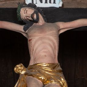 Zdjęcie nr 1: Krucyfiks z rzeźbą Chrystusa w typie Christo morte. Ciało zawieszone na niemal wyprostowanych ramionach; głowa silnie przechylona na prawe ramię; nogi delikatnie ugięte w kolanach, stopy przebite jednym gwoździem w układzie prawa na lewą. Twarz podłużna z zamkniętymi oczami i długim nosem, okolona krótkim zarostem. Na głowie zielona korona cierniowa. Włosy długie, czarne, opadające na prawe ramię oraz na plecy, silnie zawinięte do tyłu w drobne pukle. Klatka piersiowa szeroka z delikatnie, łukowato zaznaczoną linią żeber i mostka. Perizonium przewiązane na prawym boku w supeł z prawym końcem zwisającym wzdłuż prawego uda. Nad głową Chrystusa znajduje się biały titulus z napisem „INRI” w polu. Polichromia naturalistyczna w odsłoniętych partiach ciała z zaznaczonymi śladami męki, perizonium złocone. Krzyż o szerokich ramionach, prosty, malowany na czarno. 