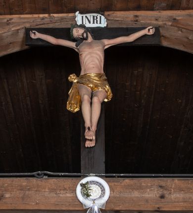 Zdjęcie nr 2: Krucyfiks z rzeźbą Chrystusa w typie Christo morte. Ciało zawieszone na niemal wyprostowanych ramionach; głowa silnie przechylona na prawe ramię; nogi delikatnie ugięte w kolanach, stopy przebite jednym gwoździem w układzie prawa na lewą. Twarz podłużna z zamkniętymi oczami i długim nosem, okolona krótkim zarostem. Na głowie zielona korona cierniowa. Włosy długie, czarne, opadające na prawe ramię oraz na plecy, silnie zawinięte do tyłu w drobne pukle. Klatka piersiowa szeroka z delikatnie, łukowato zaznaczoną linią żeber i mostka. Perizonium przewiązane na prawym boku w supeł z prawym końcem zwisającym wzdłuż prawego uda. Nad głową Chrystusa znajduje się biały titulus z napisem „INRI” w polu. Polichromia naturalistyczna w odsłoniętych partiach ciała z zaznaczonymi śladami męki, perizonium złocone. Krzyż o szerokich ramionach, prosty, malowany na czarno. 