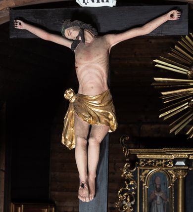 Zdjęcie nr 5: Krucyfiks z rzeźbą Chrystusa w typie Christo morte. Ciało zawieszone na niemal wyprostowanych ramionach; głowa silnie przechylona na prawe ramię; nogi delikatnie ugięte w kolanach, stopy przebite jednym gwoździem w układzie prawa na lewą. Twarz podłużna z zamkniętymi oczami i długim nosem, okolona krótkim zarostem. Na głowie zielona korona cierniowa. Włosy długie, czarne, opadające na prawe ramię oraz na plecy, silnie zawinięte do tyłu w drobne pukle. Klatka piersiowa szeroka z delikatnie, łukowato zaznaczoną linią żeber i mostka. Perizonium przewiązane na prawym boku w supeł z prawym końcem zwisającym wzdłuż prawego uda. Nad głową Chrystusa znajduje się biały titulus z napisem „INRI” w polu. Polichromia naturalistyczna w odsłoniętych partiach ciała z zaznaczonymi śladami męki, perizonium złocone. Krzyż o szerokich ramionach, prosty, malowany na czarno. 