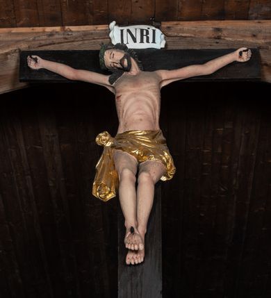 Zdjęcie nr 3: Krucyfiks z rzeźbą Chrystusa w typie Christo morte. Ciało zawieszone na niemal wyprostowanych ramionach; głowa silnie przechylona na prawe ramię; nogi delikatnie ugięte w kolanach, stopy przebite jednym gwoździem w układzie prawa na lewą. Twarz podłużna z zamkniętymi oczami i długim nosem, okolona krótkim zarostem. Na głowie zielona korona cierniowa. Włosy długie, czarne, opadające na prawe ramię oraz na plecy, silnie zawinięte do tyłu w drobne pukle. Klatka piersiowa szeroka z delikatnie, łukowato zaznaczoną linią żeber i mostka. Perizonium przewiązane na prawym boku w supeł z prawym końcem zwisającym wzdłuż prawego uda. Nad głową Chrystusa znajduje się biały titulus z napisem „INRI” w polu. Polichromia naturalistyczna w odsłoniętych partiach ciała z zaznaczonymi śladami męki, perizonium złocone. Krzyż o szerokich ramionach, prosty, malowany na czarno. 