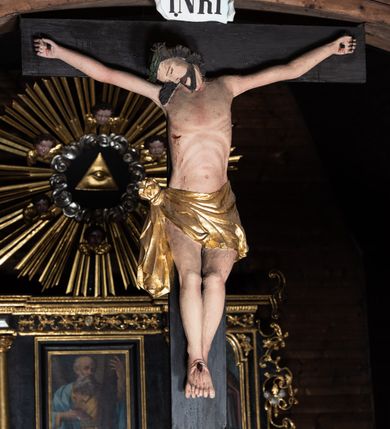 Zdjęcie nr 4: Krucyfiks z rzeźbą Chrystusa w typie Christo morte. Ciało zawieszone na niemal wyprostowanych ramionach; głowa silnie przechylona na prawe ramię; nogi delikatnie ugięte w kolanach, stopy przebite jednym gwoździem w układzie prawa na lewą. Twarz podłużna z zamkniętymi oczami i długim nosem, okolona krótkim zarostem. Na głowie zielona korona cierniowa. Włosy długie, czarne, opadające na prawe ramię oraz na plecy, silnie zawinięte do tyłu w drobne pukle. Klatka piersiowa szeroka z delikatnie, łukowato zaznaczoną linią żeber i mostka. Perizonium przewiązane na prawym boku w supeł z prawym końcem zwisającym wzdłuż prawego uda. Nad głową Chrystusa znajduje się biały titulus z napisem „INRI” w polu. Polichromia naturalistyczna w odsłoniętych partiach ciała z zaznaczonymi śladami męki, perizonium złocone. Krzyż o szerokich ramionach, prosty, malowany na czarno. 