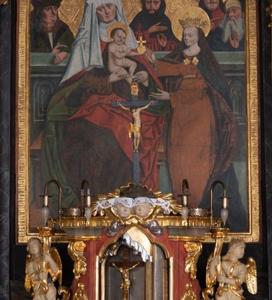 Zdjęcie nr 2: Obraz w kształcie stojącego prostokąta, ujęty profilowaną, złoconą ramą z gotycką inskrypcją. Kompozycja dwuplanowa, wieloosobowa. Na pierwszym planie ukazane postaci św. Anny zasiadającej na drewnianej ławie z Dzieciątkiem Jezus na kolanach i stojącej z prawej strony kompozycji Marii. Święta Anna ukazana jako dojrzała kobieta zwrócona w trzech czwartych w lewo z głową skierowaną nieznacznie w prawo. Rękami przytrzymuje nóżki Dzieciątka siedzącego na jej kolanach. Twarz św. Anny owalna, szeroka, z wydatnymi kośćmi policzkowymi, długim nosem i wąskimi ustami. Święta ubrana jest w brązową suknię z długimi rękawami, czerwony obficie fałdowany płaszcz z zieloną podszewką i białym kołnierzem. Na głowie ma białą chustę zasłaniającą czoło i opadającą po bokach na ramiona. Wokół głowy św. Anny znajduje się złoty nimb. Dzieciątko nagie, ukazany w pozycji siedzącej z zwrócone w lewo, w kierunku Marii. Prawą ręką błogosławi, na lewej dłoni trzyma złote jabłko zwieńczone krzyżem. Jezus o okrągłej twarzy z wysokim czołem i krótkimi, brązowymi włosami; wokół głowy ma złoty nimb. Ukazana z prawej trony kompozycji Maria została przedstawiona jako młoda kobieta o długich, brązowych, lekko falowanych włosach zwracająca się całym ciałem do Dzieciątka Jezus, do którego wyciąga dłonie. Twarz Marii młodzieńcza, z długim, wąskim nosem i małymi ustami, wzrok skierowany w stronę Dzieciątka. Policzki zarumienione, wysokie czoło, włosy opadają na ramiona i plecy. Maria jest ubrana w jasnobrązową suknię z długimi rękawami, przepasaną pod biustem. Na ramiona ma narzucony ciemnoniebieski płaszcz z ciemnobrązową podszewką dekorowaną kwiatowym ornamentem spięty pod szyją złotą broszą. Na głowie Marii złocona i kameryzowana korona w typie otwartym, wokół głowy złoty nimb. Na drugim planie, za kamiennym parapetem dekorowanym maswerkiem ukazane postaci czterech mężczyzn – św. Józefa oraz trzech mężów św. Anny: Joachima, Kleofasa i Salomasa. Z lewej strony kompozycji ukazany długowłosy mężczyzna w czerwono-zielonym kapeluszu, zielonym płaszczu z futrzanym kołnierzem i białej szacie spodniej ze złotą lamówką pod szyją. Mężczyzna o twarzy pozbawionej zarostu, długich i ciemnych włosach zwraca twarz w lewo. Jego lewa dłoń spoczywa na kamiennym parapecie, drugą wskazuje w lewo. Kolejny mężczyzna ukazany jest frontalnie. Ma krótkie, ciemne włosy i gęstą ciemną brodę rozdwajającą się na końcu. Ciemne oczy skierowane są wprost na widza. Prawą dłonią wskazuje w lewą stronę. Ubrany jest w obszerną czerwoną suknię i ciemnogranatowy płaszcz z kapturem. Kolejni dwaj mężczyźni zostali ukazani z prawej strony kompozycji. Pierwszy z nich przedstawiony w czerwonym, zapinanym na guziki kaftanie i białym turbanie o szerokiej, owalnej twarzy, długich, prostych włosach i niewielkim wąsie. Wskazuje on w lewo na ostatniego z mężczyzn, zapewne wyobrażającego św. Józefa. Postać ma długą, siwą brodę rozdwajającą się na końcu oraz wydatny nos. Ubrany jest w granatowe szaty oraz zielono-żółty kapelusz. Lewą dłoń unosi w geście pozdrowienia. Tło złocone, wytłaczane w motyw skośnej kraty z wpisanymi w nią stylizowanymi motywami roślinnymi.