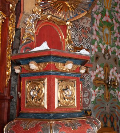 Zdjęcie nr 4: Ambona zawieszona przy północnej ścianie prezbiterium z wejściem od strony zakrystii. Kosz na planie sześcioboku z dodanym uskokiem po prawej stronie, od dołu ujęty jest wydatnym półwałkiem. Podstawa stożkowata, zwężająca się ku dołowi, zakończona zwornikiem z wolutami. Kosz został zamknięty belkowaniem z wydatnym gzymsem. Zaplecek jest prostokątny, ujęty spływami wolutowymi. W jego polu prostokątna płycina zamknięta łukiem nadwieszonym. Skośnie powyżej został nadwieszony wieloboczny baldachim z glorią promienistą w podniebiu z symbolem oka Opatrzności Bożej; w polu zaplecka wycięto drzwi. Boki kosza artykułowane płycinami wypełnionymi wolutowo zwiniętym liściem akantu z rozetą. Podstawa dekorowana aplikowanymi, wywiniętymi liśćmi akantu. W partii fryzu rzeźbione uskrzydlone główki anielskie. Zaplecek oraz baldachim oplecione płaskorzeźbioną tkaniną z supłami, której podwiązane końce opadają na woluty zaplecka, formując kaskadowe fałdy. Dolna partia kosza oraz woluty zaplecka dekorowane groszkowaniem. Całość struktury malowana w kolorze brązowo-czerwonym, profilowania marmoryzowane w odcieniu ciemnoniebieskim, detal architektoniczny pozłacany i posrebrzany.