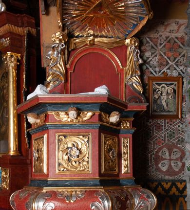 Zdjęcie nr 2: Ambona zawieszona przy północnej ścianie prezbiterium z wejściem od strony zakrystii. Kosz na planie sześcioboku z dodanym uskokiem po prawej stronie, od dołu ujęty jest wydatnym półwałkiem. Podstawa stożkowata, zwężająca się ku dołowi, zakończona zwornikiem z wolutami. Kosz został zamknięty belkowaniem z wydatnym gzymsem. Zaplecek jest prostokątny, ujęty spływami wolutowymi. W jego polu prostokątna płycina zamknięta łukiem nadwieszonym. Skośnie powyżej został nadwieszony wieloboczny baldachim z glorią promienistą w podniebiu z symbolem oka Opatrzności Bożej; w polu zaplecka wycięto drzwi. Boki kosza artykułowane płycinami wypełnionymi wolutowo zwiniętym liściem akantu z rozetą. Podstawa dekorowana aplikowanymi, wywiniętymi liśćmi akantu. W partii fryzu rzeźbione uskrzydlone główki anielskie. Zaplecek oraz baldachim oplecione płaskorzeźbioną tkaniną z supłami, której podwiązane końce opadają na woluty zaplecka, formując kaskadowe fałdy. Dolna partia kosza oraz woluty zaplecka dekorowane groszkowaniem. Całość struktury malowana w kolorze brązowo-czerwonym, profilowania marmoryzowane w odcieniu ciemnoniebieskim, detal architektoniczny pozłacany i posrebrzany.