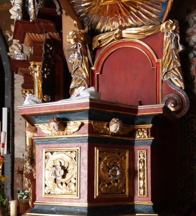Zdjęcie nr 3: Ambona zawieszona przy północnej ścianie prezbiterium z wejściem od strony zakrystii. Kosz na planie sześcioboku z dodanym uskokiem po prawej stronie, od dołu ujęty jest wydatnym półwałkiem. Podstawa stożkowata, zwężająca się ku dołowi, zakończona zwornikiem z wolutami. Kosz został zamknięty belkowaniem z wydatnym gzymsem. Zaplecek jest prostokątny, ujęty spływami wolutowymi. W jego polu prostokątna płycina zamknięta łukiem nadwieszonym. Skośnie powyżej został nadwieszony wieloboczny baldachim z glorią promienistą w podniebiu z symbolem oka Opatrzności Bożej; w polu zaplecka wycięto drzwi. Boki kosza artykułowane płycinami wypełnionymi wolutowo zwiniętym liściem akantu z rozetą. Podstawa dekorowana aplikowanymi, wywiniętymi liśćmi akantu. W partii fryzu rzeźbione uskrzydlone główki anielskie. Zaplecek oraz baldachim oplecione płaskorzeźbioną tkaniną z supłami, której podwiązane końce opadają na woluty zaplecka, formując kaskadowe fałdy. Dolna partia kosza oraz woluty zaplecka dekorowane groszkowaniem. Całość struktury malowana w kolorze brązowo-czerwonym, profilowania marmoryzowane w odcieniu ciemnoniebieskim, detal architektoniczny pozłacany i posrebrzany.