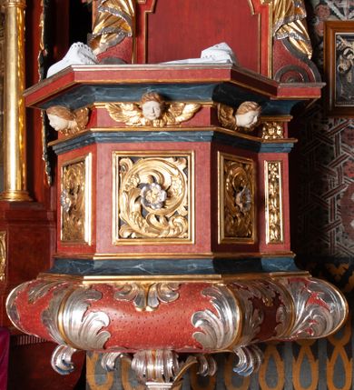 Zdjęcie nr 1: Ambona zawieszona przy północnej ścianie prezbiterium z wejściem od strony zakrystii. Kosz na planie sześcioboku z dodanym uskokiem po prawej stronie, od dołu ujęty jest wydatnym półwałkiem. Podstawa stożkowata, zwężająca się ku dołowi, zakończona zwornikiem z wolutami. Kosz został zamknięty belkowaniem z wydatnym gzymsem. Zaplecek jest prostokątny, ujęty spływami wolutowymi. W jego polu prostokątna płycina zamknięta łukiem nadwieszonym. Skośnie powyżej został nadwieszony wieloboczny baldachim z glorią promienistą w podniebiu z symbolem oka Opatrzności Bożej; w polu zaplecka wycięto drzwi. Boki kosza artykułowane płycinami wypełnionymi wolutowo zwiniętym liściem akantu z rozetą. Podstawa dekorowana aplikowanymi, wywiniętymi liśćmi akantu. W partii fryzu rzeźbione uskrzydlone główki anielskie. Zaplecek oraz baldachim oplecione płaskorzeźbioną tkaniną z supłami, której podwiązane końce opadają na woluty zaplecka, formując kaskadowe fałdy. Dolna partia kosza oraz woluty zaplecka dekorowane groszkowaniem. Całość struktury malowana w kolorze brązowo-czerwonym, profilowania marmoryzowane w odcieniu ciemnoniebieskim, detal architektoniczny pozłacany i posrebrzany.