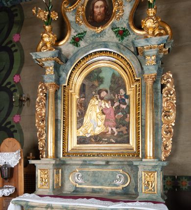 Zdjęcie nr 2: Nastawa ołtarzowa drewniana, architektoniczna, jednoosiowa, jednokondygnacyjna ze zwieńczeniem. Mensa w typie sarkofagowym, wklęsło-wypukła, zwężający się ku dołowi, zamknięta gzymsem; naroża ujęte fragmentami rocaille'u, na froncie rocaille’owy kartusz; portatyl kamienny. Retabulum posadowione na cokole zryzalitowanym po bokach, zamkniętym silnie wysuniętym gzymsem; ujęte parą kolumn o kapitelach korynckich dźwigających imposty, których fryz przebiega przez całą szerokość retabulum, przyjmując nad polem środkowym formę łuku półkolistego nadwieszonego. W polu środkowym znajduje się nisza w formie kwadratu zamkniętego łukiem półkolistym nadwieszonym z płaskorzeźbioną sceną Świętej Rodziny w warsztacie, ujętego trzema ramami. Zwieńczenie ma formę zbliżoną do trapezu. Jest flankowane spływami wolutowymi i zamknięte baldachimem z profilowanym cokołem. W polu medalion otoczony profilowaną ramą z obrazem „Veraikon”. Cokoły kolumn dekorowane z trzech stron płycinami z ornamentem roślinnym; w polu predelli płycina w formie leżącego prostokąta o wypukłych ściankach bocznych z ornamentem roślinnym. Kondygnacja ujęta uszami utworzonymi z mięsistego akantu. Na fryzie rozety; na impostach rokokowe wazony z dużymi kwiatami słonecznika, róż i lilii, rama obrazu zwieńczenia dekorowana ornamentem roślinnym; u spodu akant z rozetami. Baldachim z podwieszonym lambrekinem; na jego cokole rokokowy wazon z dużymi kwiatami słonecznika, róż i lilii. Struktura marmoryzowana w kolorze szaro-niebieskim; ornamentyka, profile i kolumny złocone i srebrzone.