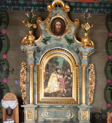 Zdjęcie nr 1: Nastawa ołtarzowa drewniana, architektoniczna, jednoosiowa, jednokondygnacyjna ze zwieńczeniem. Mensa w typie sarkofagowym, wklęsło-wypukła, zwężający się ku dołowi, zamknięta gzymsem; naroża ujęte fragmentami rocaille'u, na froncie rocaille’owy kartusz; portatyl kamienny. Retabulum posadowione na cokole zryzalitowanym po bokach, zamkniętym silnie wysuniętym gzymsem; ujęte parą kolumn o kapitelach korynckich dźwigających imposty, których fryz przebiega przez całą szerokość retabulum, przyjmując nad polem środkowym formę łuku półkolistego nadwieszonego. W polu środkowym znajduje się nisza w formie kwadratu zamkniętego łukiem półkolistym nadwieszonym z płaskorzeźbioną sceną Świętej Rodziny w warsztacie, ujętego trzema ramami. Zwieńczenie ma formę zbliżoną do trapezu. Jest flankowane spływami wolutowymi i zamknięte baldachimem z profilowanym cokołem. W polu medalion otoczony profilowaną ramą z obrazem „Veraikon”. Cokoły kolumn dekorowane z trzech stron płycinami z ornamentem roślinnym; w polu predelli płycina w formie leżącego prostokąta o wypukłych ściankach bocznych z ornamentem roślinnym. Kondygnacja ujęta uszami utworzonymi z mięsistego akantu. Na fryzie rozety; na impostach rokokowe wazony z dużymi kwiatami słonecznika, róż i lilii, rama obrazu zwieńczenia dekorowana ornamentem roślinnym; u spodu akant z rozetami. Baldachim z podwieszonym lambrekinem; na jego cokole rokokowy wazon z dużymi kwiatami słonecznika, róż i lilii. Struktura marmoryzowana w kolorze szaro-niebieskim; ornamentyka, profile i kolumny złocone i srebrzone.