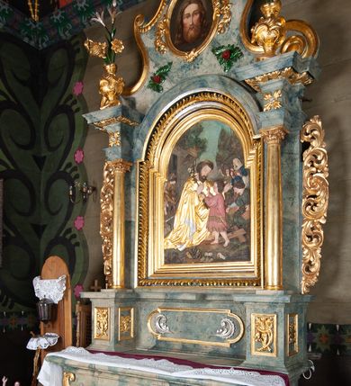 Zdjęcie nr 3: Nastawa ołtarzowa drewniana, architektoniczna, jednoosiowa, jednokondygnacyjna ze zwieńczeniem. Mensa w typie sarkofagowym, wklęsło-wypukła, zwężający się ku dołowi, zamknięta gzymsem; naroża ujęte fragmentami rocaille&#039;u, na froncie rocaille’owy kartusz; portatyl kamienny. Retabulum posadowione na cokole zryzalitowanym po bokach, zamkniętym silnie wysuniętym gzymsem; ujęte parą kolumn o kapitelach korynckich dźwigających imposty, których fryz przebiega przez całą szerokość retabulum, przyjmując nad polem środkowym formę łuku półkolistego nadwieszonego. W polu środkowym znajduje się nisza w formie kwadratu zamkniętego łukiem półkolistym nadwieszonym z płaskorzeźbioną sceną Świętej Rodziny w warsztacie, ujętego trzema ramami. Zwieńczenie ma formę zbliżoną do trapezu. Jest flankowane spływami wolutowymi i zamknięte baldachimem z profilowanym cokołem. W polu medalion otoczony profilowaną ramą z obrazem „Veraikon”. Cokoły kolumn dekorowane z trzech stron płycinami z ornamentem roślinnym; w polu predelli płycina w formie leżącego prostokąta o wypukłych ściankach bocznych z ornamentem roślinnym. Kondygnacja ujęta uszami utworzonymi z mięsistego akantu. Na fryzie rozety; na impostach rokokowe wazony z dużymi kwiatami słonecznika, róż i lilii, rama obrazu zwieńczenia dekorowana ornamentem roślinnym; u spodu akant z rozetami. Baldachim z podwieszonym lambrekinem; na jego cokole rokokowy wazon z dużymi kwiatami słonecznika, róż i lilii. Struktura marmoryzowana w kolorze szaro-niebieskim; ornamentyka, profile i kolumny złocone i srebrzone.