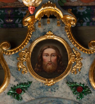 Zdjęcie nr 6: Nastawa ołtarzowa drewniana, architektoniczna, jednoosiowa, jednokondygnacyjna ze zwieńczeniem. Mensa w typie sarkofagowym, wklęsło-wypukła, zwężający się ku dołowi, zamknięta gzymsem; naroża ujęte fragmentami rocaille'u, na froncie rocaille’owy kartusz; portatyl kamienny. Retabulum posadowione na cokole zryzalitowanym po bokach, zamkniętym silnie wysuniętym gzymsem; ujęte parą kolumn o kapitelach korynckich dźwigających imposty, których fryz przebiega przez całą szerokość retabulum, przyjmując nad polem środkowym formę łuku półkolistego nadwieszonego. W polu środkowym znajduje się nisza w formie kwadratu zamkniętego łukiem półkolistym nadwieszonym z płaskorzeźbioną sceną Świętej Rodziny w warsztacie, ujętego trzema ramami. Zwieńczenie ma formę zbliżoną do trapezu. Jest flankowane spływami wolutowymi i zamknięte baldachimem z profilowanym cokołem. W polu medalion otoczony profilowaną ramą z obrazem „Veraikon”. Cokoły kolumn dekorowane z trzech stron płycinami z ornamentem roślinnym; w polu predelli płycina w formie leżącego prostokąta o wypukłych ściankach bocznych z ornamentem roślinnym. Kondygnacja ujęta uszami utworzonymi z mięsistego akantu. Na fryzie rozety; na impostach rokokowe wazony z dużymi kwiatami słonecznika, róż i lilii, rama obrazu zwieńczenia dekorowana ornamentem roślinnym; u spodu akant z rozetami. Baldachim z podwieszonym lambrekinem; na jego cokole rokokowy wazon z dużymi kwiatami słonecznika, róż i lilii. Struktura marmoryzowana w kolorze szaro-niebieskim; ornamentyka, profile i kolumny złocone i srebrzone.