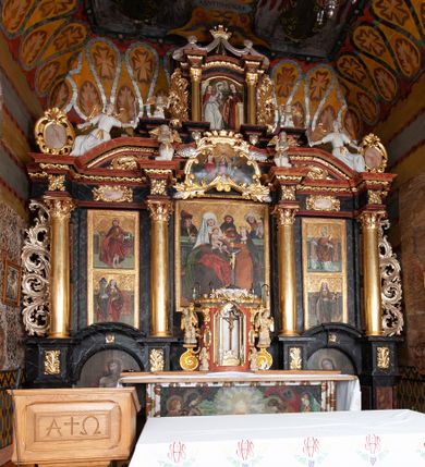 Zdjęcie nr 13: Ołtarz trójosiowy, dwukondygnacyjny z bramkami o formach architektonicznych, uszaty. Mensa murowana, prostopadłościenna z drewnianym antepedium ujętym profilowaną ramą z malowaną dekoracją roślinną. W polu malowane na płótnie przedstawienie adoracji Najświętszego Sakramentu. Tabernakulum w formie prostopadłościanu, o wypukłej ściance frontowej, ujętej pilastrami o kapitelach w formie akantowych konsol podtrzymujących pełne belkowanie. Na trzonach owalne lustra w akantowych ramach. W zwieńczeniu dwie uskrzydlone główki anielskie. Na metalowych drzwiczkach krucyfiks oraz kielich z hostią w otoczeniu winnych gron i kłosów (na odwrociu). Naroża tabernakulum flankuje para aniołów ustawionych na wolutowych konsolach. Nastawa umieszczona na wysokim cokole z bramkami mieszczącymi obrazy Chrystusa w typie Ecce Homo (z lewej) oraz Matki Boskiej Bolesnej (z prawej). Cokół artykułowany wysokimi postumentami, na których ustawione są kolumny. W polach postumentów złocona dekoracja akantowa. Kondygnacja pierwsza trójosiowa, artykułowana złoconymi kolumnami o korynckich kapitelach, ujęta srebrzonymi akantowymi uszami (suchy akant). Kolumny dźwigają pełne belkowanie, wyłamane na osiach podpór oraz odcinki przyczółka, na których siedzą figury aniołów w białych szatach. Dodatkowo, w osiach kolumn umieszczone mniejsze figury klęczących aniołów (osie wewnętrzne) oraz marmoryzowane, owalne kartusze ujęte złoconymi ramami z ornamentem małżowinowo-chrząstkowym (osie zewnętrzne). W partii fryzu złocone rozetki oraz marmoryzowane, owalne kartusze ujęte złoconym akantem. Gzyms profilowany, dekorowany dodatkowo złoconym kimationem. W polu środkowym prostokątny obraz przedstawiający Rodzinę Marii. Na zasuwie umieszczony obraz z wizerunkiem św. Katarzyny Aleksandryjskiej. Powyżej, w osi ołtarza obraz ze sceną Koronacji Marii, o nieregularnym kształcie w akantowej ramie, zwieńczonej koroną. W osiach bocznych umieszczone skrzydła boczne dawnego, gotyckiego tryptyku z wizerunkami św. Jana Chrzciciela i św. Barbary (z lewej) oraz św. Stanisława biskupa i św. Katarzyny Aleksandryjskiej (z prawej). Zwieńczenie ołtarza w formie edikuli ujętej złoconymi, korynckimi kolumnami oraz złoconymi, akantowymi uszami. Po bokach niewielkie figury klęczących aniołów. Podpory dźwigają wyłamane belkowanie dekorowane kimationem i rozetkami, łukowato wyginające się w osi ołtarza. W polu obraz w kształcie stojącego prostokąta zamkniętego łukiem półkolistym ze sceną Zwiastowania. Całość zwieńczona srebrzonym baldachimem z podwieszonej tkaniny przewiązanej złotym sznurem ze złoconą koroną umieszczoną na szczycie. Struktura ołtarza marmoryzowana w odcieniu ciemnoszarym, profile i płyciny na filarach marmoryzowane w kolorze bordowym, ornamentyka złocona i posrebrzana.