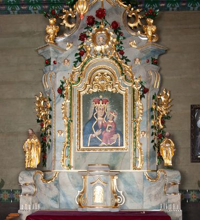 Zdjęcie nr 9: Nastawa ołtarzowa drewniana, architektoniczna, jednoosiowa, jednokondygnacyjna ze zwieńczeniem. Mensa w typie sarkofagowym, wklęsło-wypukła, zwężająca się ku dołowi, zamknięta gzymsem; naroża ujęte fragmentami rocaille&#039;u, na froncie rocaille’owy kartusz; portatyl z marmuru dębnickiego. Na mensie ustawiono tabernakulum w formie prostopadłościennej skrzynki, o wypukłej ściance frontowej, która jest ujęta w narożach ustawionymi pod kątem pilastrami na cokołach, których trzony przyjmują formę spływów wolutowych. Podpory dźwigają gzyms, opasujący całą skrzynkę, wyłamany na ich osi. Drzwiczki w formie stojącego prostokąta zamkniętego łukiem nadwieszonym, w polu nisza z płaskorzeźbionym przedstawieniem kielicha z hostią w glorii. Drzwiczki zostały ujęte płyciną w formie stojącego prostokąta zamkniętego łukiem kotarowym dwudzielnym. Na tabernakulum tron wystawienia w postaci cokołu. Nastawa ołtarzowa została posadowiona na wysokim cokole, przyjmującym po bokach formę wolut stanowiących podstawę dla figur św. Piotra z prawej i św. Pawła z lewej strony. Jest ujęta parą pilastrów ustawionych pod katem względem lica, których trzony u góry przechodzą w woluty. Podpory dźwigają imposty. W polu środkowym płycina w formie stojącego prostokąta zamkniętego łukiem półkolistym nadwieszonym z uskokiem, w której znajduje się mniejsza nisza o tej samej formie, ujęta ramą, wypełniona obrazem Matki Boskiej z Dzieciątkiem. Zwieńczenie w formie zbliżonej do trapezu, ujętego podwójnymi spływami wolutowymi, zamkniętego falistym gzymsem. Całość wieńczy rzeźbiona gołębica Ducha Świętego w glorii promienistej. Trzony pilastrów nastawy dekorowane zwisami z różanych gałązek. Płycina pola środkowego ujęta rocaille&#039;em, który ponad łukiem, w polu zwieńczenia, tworzy medalion z uskrzydloną główką anielską, otoczony dużymi różanymi gałązkami; w polu środkowym cztery uskrzydlone główki anielskie. Kondygnacja flankowana uszami utworzonymi z płomienistego rocaille&#039;u i różanych gałązek. Na belkowaniu dwa rokokowe wazony z różami. Zwieńczenie ujęte falbanką, rocaille&#039;em i różanymi gałązkami. Struktura w całości marmoryzowana w kolorze szaroniebieskim, ornamentyki i profile złocone i srebrzone, ukwiecone gałązki laserunkowe.