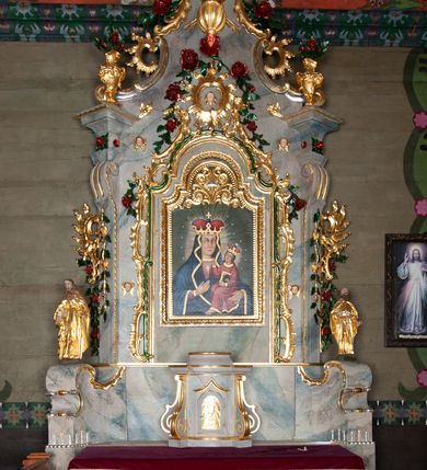 Zdjęcie nr 2: Nastawa ołtarzowa drewniana, architektoniczna, jednoosiowa, jednokondygnacyjna ze zwieńczeniem. Mensa w typie sarkofagowym, wklęsło-wypukła, zwężająca się ku dołowi, zamknięta gzymsem; naroża ujęte fragmentami rocaille'u, na froncie rocaille’owy kartusz; portatyl z marmuru dębnickiego. Na mensie ustawiono tabernakulum w formie prostopadłościennej skrzynki, o wypukłej ściance frontowej, która jest ujęta w narożach ustawionymi pod kątem pilastrami na cokołach, których trzony przyjmują formę spływów wolutowych. Podpory dźwigają gzyms, opasujący całą skrzynkę, wyłamany na ich osi. Drzwiczki w formie stojącego prostokąta zamkniętego łukiem nadwieszonym, w polu nisza z płaskorzeźbionym przedstawieniem kielicha z hostią w glorii. Drzwiczki zostały ujęte płyciną w formie stojącego prostokąta zamkniętego łukiem kotarowym dwudzielnym. Na tabernakulum tron wystawienia w postaci cokołu. Nastawa ołtarzowa została posadowiona na wysokim cokole, przyjmującym po bokach formę wolut stanowiących podstawę dla figur św. Piotra z prawej i św. Pawła z lewej strony. Jest ujęta parą pilastrów ustawionych pod katem względem lica, których trzony u góry przechodzą w woluty. Podpory dźwigają imposty. W polu środkowym płycina w formie stojącego prostokąta zamkniętego łukiem półkolistym nadwieszonym z uskokiem, w której znajduje się mniejsza nisza o tej samej formie, ujęta ramą, wypełniona obrazem Matki Boskiej z Dzieciątkiem. Zwieńczenie w formie zbliżonej do trapezu, ujętego podwójnymi spływami wolutowymi, zamkniętego falistym gzymsem. Całość wieńczy rzeźbiona gołębica Ducha Świętego w glorii promienistej. Trzony pilastrów nastawy dekorowane zwisami z różanych gałązek. Płycina pola środkowego ujęta rocaille'em, który ponad łukiem, w polu zwieńczenia, tworzy medalion z uskrzydloną główką anielską, otoczony dużymi różanymi gałązkami; w polu środkowym cztery uskrzydlone główki anielskie. Kondygnacja flankowana uszami utworzonymi z płomienistego rocaille'u i różanych gałązek. Na belkowaniu dwa rokokowe wazony z różami. Zwieńczenie ujęte falbanką, rocaille'em i różanymi gałązkami. Struktura w całości marmoryzowana w kolorze szaroniebieskim, ornamentyki i profile złocone i srebrzone, ukwiecone gałązki laserunkowe.
