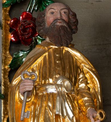 Zdjęcie nr 1: Rzeźba ścięta z tyłu została ustawiona na podstawie w formie kostki o ściętych narożach. Figura jest zwrócona frontalnie, w delikatnym kontrapoście. Święty ma obie ręce, ugięte, uniesione, w dłoniach trzyma klucze – w prawej srebrny, w lewej złoty. Twarz ma pociągłą, szczupłą, o płytko osadzonych oczach, krótkim, prostym nosie i okoloną falowaną, opadającą na pierś brodą. Włosy wywinięte w loki, gęste, sięgają karku. Święty Piotr jest ubrany w złotą tunikę z długimi, wąskimi rękawami, przepasaną w talii srebrną wstęgą, zawiązaną z przodu na węzeł, drapowaną w liczne, równoległe fałdki oraz złoty płaszcz zapięty pod szyją, opadający na ramiona i delikatnie drapowany. Polichromia naturalistyczna w odsłoniętych partiach ciała. 