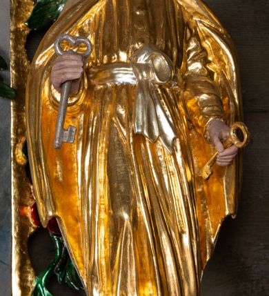 Zdjęcie nr 2: Rzeźba ścięta z tyłu została ustawiona na podstawie w formie kostki o ściętych narożach. Figura jest zwrócona frontalnie, w delikatnym kontrapoście. Święty ma obie ręce, ugięte, uniesione, w dłoniach trzyma klucze – w prawej srebrny, w lewej złoty. Twarz ma pociągłą, szczupłą, o płytko osadzonych oczach, krótkim, prostym nosie i okoloną falowaną, opadającą na pierś brodą. Włosy wywinięte w loki, gęste, sięgają karku. Święty Piotr jest ubrany w złotą tunikę z długimi, wąskimi rękawami, przepasaną w talii srebrną wstęgą, zawiązaną z przodu na węzeł, drapowaną w liczne, równoległe fałdki oraz złoty płaszcz zapięty pod szyją, opadający na ramiona i delikatnie drapowany. Polichromia naturalistyczna w odsłoniętych partiach ciała. 