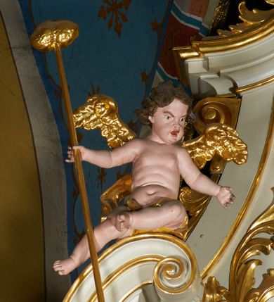 Zdjęcie nr 11: Ołtarz prostopadłościenny z mensą. Antepedium w formie leżącego prostokąta ujętego drewnianą, profilowaną ramą z przedstawieniem Mandylionu i narzędziami Męki Pańskiej na tle wypełnionym ornamentem roślinnym. W centrum, u dołu dwie tarcze herbowe z inicjałami: „TR” i „W”.
Nastawa ołtarzowa architektoniczna, jednoosiowa z bramkami, jednokondygnacyjna ze zwieńczeniem, ustawiona na dwustrefowym cokole. Ujęta dwiema parami kolumn o kapitelach korynckich, ustawionych uskokowo na cokołach zdobionych ornamentem rocaille, dźwigających pełne belkowanie wyłamane na ich osi, nad polem głównym przerwany architraw i fryz oraz gzyms przyjmujący kształt łuku odcinkowego. Na belkowaniu wewnętrznej pary kolumn przerwany wolutowy przyczółek z siedzącymi na nim puttami z narzędziami Męki Pańskiej. W polu głównym wnęka o wykroju prostokąta stojącego zamkniętego łukiem odcinkowym nadwieszonym, w złotej ramie dekorowanej rocaille, w której znajduje się krucyfiks na tle czerwonego atłasu obwieszonego wotami. Powyżej, w partii belkowania rocaillowy kartusz z napisem: „HOC OPUS / NOSTRAE / SALUTIS”. Ołtarz ujęty ażurowymi uszami utworzonymi z ornamentu rocaille.  Zwieńczenie ujęte spływami wolutowymi, zamknięte dwiema skierowanymi do środka wolutami, dekorowane ornamentem rocaille. W polu płycina w formie stojącego owalu wyłożona czerwonym atłasem, ujęta profilowaną ramą z rzeźbą gołębicy Ducha Świętego na tle obłoków w glorii promienistej, flankowana przez akant przechodzący w woluty oraz przez kwiatowe festony z wstęgą. Bramki o wykroju stojącego prostokąta, zamkniętego łukiem odcinkowym nadwieszonym z drzwiczkami, zakończone konsolami ujętymi spływami wolutowymi, stanowiącymi podstawy dla figur św. Michała Archanioła i św. Wacława. Struktura ołtarza malowana na kolor biały, z pozłacaną ornamentyką profilami i kolumnami.

