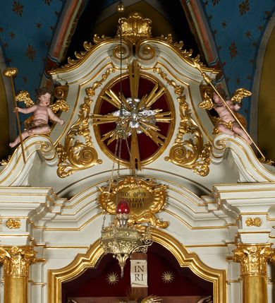 Zdjęcie nr 26: Ołtarz prostopadłościenny z mensą. Antepedium w formie leżącego prostokąta ujętego drewnianą, profilowaną ramą z przedstawieniem Mandylionu i narzędziami Męki Pańskiej na tle wypełnionym ornamentem roślinnym. W centrum, u dołu dwie tarcze herbowe z inicjałami: „TR” i „W”.
Nastawa ołtarzowa architektoniczna, jednoosiowa z bramkami, jednokondygnacyjna ze zwieńczeniem, ustawiona na dwustrefowym cokole. Ujęta dwiema parami kolumn o kapitelach korynckich, ustawionych uskokowo na cokołach zdobionych ornamentem rocaille, dźwigających pełne belkowanie wyłamane na ich osi, nad polem głównym przerwany architraw i fryz oraz gzyms przyjmujący kształt łuku odcinkowego. Na belkowaniu wewnętrznej pary kolumn przerwany wolutowy przyczółek z siedzącymi na nim puttami z narzędziami Męki Pańskiej. W polu głównym wnęka o wykroju prostokąta stojącego zamkniętego łukiem odcinkowym nadwieszonym, w złotej ramie dekorowanej rocaille, w której znajduje się krucyfiks na tle czerwonego atłasu obwieszonego wotami. Powyżej, w partii belkowania rocaillowy kartusz z napisem: „HOC OPUS / NOSTRAE / SALUTIS”. Ołtarz ujęty ażurowymi uszami utworzonymi z ornamentu rocaille.  Zwieńczenie ujęte spływami wolutowymi, zamknięte dwiema skierowanymi do środka wolutami, dekorowane ornamentem rocaille. W polu płycina w formie stojącego owalu wyłożona czerwonym atłasem, ujęta profilowaną ramą z rzeźbą gołębicy Ducha Świętego na tle obłoków w glorii promienistej, flankowana przez akant przechodzący w woluty oraz przez kwiatowe festony z wstęgą. Bramki o wykroju stojącego prostokąta, zamkniętego łukiem odcinkowym nadwieszonym z drzwiczkami, zakończone konsolami ujętymi spływami wolutowymi, stanowiącymi podstawy dla figur św. Michała Archanioła i św. Wacława. Struktura ołtarza malowana na kolor biały, z pozłacaną ornamentyką profilami i kolumnami.

