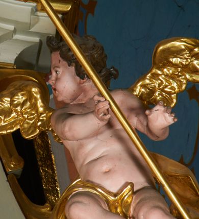 Zdjęcie nr 16: Ołtarz prostopadłościenny z mensą. Antepedium w formie leżącego prostokąta ujętego drewnianą, profilowaną ramą z przedstawieniem Mandylionu i narzędziami Męki Pańskiej na tle wypełnionym ornamentem roślinnym. W centrum, u dołu dwie tarcze herbowe z inicjałami: „TR” i „W”.
Nastawa ołtarzowa architektoniczna, jednoosiowa z bramkami, jednokondygnacyjna ze zwieńczeniem, ustawiona na dwustrefowym cokole. Ujęta dwiema parami kolumn o kapitelach korynckich, ustawionych uskokowo na cokołach zdobionych ornamentem rocaille, dźwigających pełne belkowanie wyłamane na ich osi, nad polem głównym przerwany architraw i fryz oraz gzyms przyjmujący kształt łuku odcinkowego. Na belkowaniu wewnętrznej pary kolumn przerwany wolutowy przyczółek z siedzącymi na nim puttami z narzędziami Męki Pańskiej. W polu głównym wnęka o wykroju prostokąta stojącego zamkniętego łukiem odcinkowym nadwieszonym, w złotej ramie dekorowanej rocaille, w której znajduje się krucyfiks na tle czerwonego atłasu obwieszonego wotami. Powyżej, w partii belkowania rocaillowy kartusz z napisem: „HOC OPUS / NOSTRAE / SALUTIS”. Ołtarz ujęty ażurowymi uszami utworzonymi z ornamentu rocaille.  Zwieńczenie ujęte spływami wolutowymi, zamknięte dwiema skierowanymi do środka wolutami, dekorowane ornamentem rocaille. W polu płycina w formie stojącego owalu wyłożona czerwonym atłasem, ujęta profilowaną ramą z rzeźbą gołębicy Ducha Świętego na tle obłoków w glorii promienistej, flankowana przez akant przechodzący w woluty oraz przez kwiatowe festony z wstęgą. Bramki o wykroju stojącego prostokąta, zamkniętego łukiem odcinkowym nadwieszonym z drzwiczkami, zakończone konsolami ujętymi spływami wolutowymi, stanowiącymi podstawy dla figur św. Michała Archanioła i św. Wacława. Struktura ołtarza malowana na kolor biały, z pozłacaną ornamentyką profilami i kolumnami.

