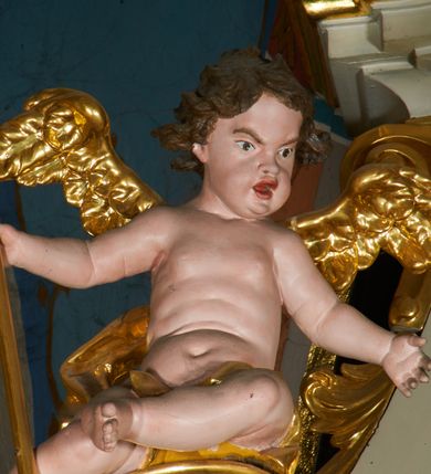 Zdjęcie nr 12: Ołtarz prostopadłościenny z mensą. Antepedium w formie leżącego prostokąta ujętego drewnianą, profilowaną ramą z przedstawieniem Mandylionu i narzędziami Męki Pańskiej na tle wypełnionym ornamentem roślinnym. W centrum, u dołu dwie tarcze herbowe z inicjałami: „TR” i „W”.
Nastawa ołtarzowa architektoniczna, jednoosiowa z bramkami, jednokondygnacyjna ze zwieńczeniem, ustawiona na dwustrefowym cokole. Ujęta dwiema parami kolumn o kapitelach korynckich, ustawionych uskokowo na cokołach zdobionych ornamentem rocaille, dźwigających pełne belkowanie wyłamane na ich osi, nad polem głównym przerwany architraw i fryz oraz gzyms przyjmujący kształt łuku odcinkowego. Na belkowaniu wewnętrznej pary kolumn przerwany wolutowy przyczółek z siedzącymi na nim puttami z narzędziami Męki Pańskiej. W polu głównym wnęka o wykroju prostokąta stojącego zamkniętego łukiem odcinkowym nadwieszonym, w złotej ramie dekorowanej rocaille, w której znajduje się krucyfiks na tle czerwonego atłasu obwieszonego wotami. Powyżej, w partii belkowania rocaillowy kartusz z napisem: „HOC OPUS / NOSTRAE / SALUTIS”. Ołtarz ujęty ażurowymi uszami utworzonymi z ornamentu rocaille.  Zwieńczenie ujęte spływami wolutowymi, zamknięte dwiema skierowanymi do środka wolutami, dekorowane ornamentem rocaille. W polu płycina w formie stojącego owalu wyłożona czerwonym atłasem, ujęta profilowaną ramą z rzeźbą gołębicy Ducha Świętego na tle obłoków w glorii promienistej, flankowana przez akant przechodzący w woluty oraz przez kwiatowe festony z wstęgą. Bramki o wykroju stojącego prostokąta, zamkniętego łukiem odcinkowym nadwieszonym z drzwiczkami, zakończone konsolami ujętymi spływami wolutowymi, stanowiącymi podstawy dla figur św. Michała Archanioła i św. Wacława. Struktura ołtarza malowana na kolor biały, z pozłacaną ornamentyką profilami i kolumnami.

