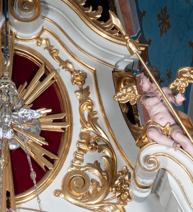 Zdjęcie nr 28: Ołtarz prostopadłościenny z mensą. Antepedium w formie leżącego prostokąta ujętego drewnianą, profilowaną ramą z przedstawieniem Mandylionu i narzędziami Męki Pańskiej na tle wypełnionym ornamentem roślinnym. W centrum, u dołu dwie tarcze herbowe z inicjałami: „TR” i „W”.
Nastawa ołtarzowa architektoniczna, jednoosiowa z bramkami, jednokondygnacyjna ze zwieńczeniem, ustawiona na dwustrefowym cokole. Ujęta dwiema parami kolumn o kapitelach korynckich, ustawionych uskokowo na cokołach zdobionych ornamentem rocaille, dźwigających pełne belkowanie wyłamane na ich osi, nad polem głównym przerwany architraw i fryz oraz gzyms przyjmujący kształt łuku odcinkowego. Na belkowaniu wewnętrznej pary kolumn przerwany wolutowy przyczółek z siedzącymi na nim puttami z narzędziami Męki Pańskiej. W polu głównym wnęka o wykroju prostokąta stojącego zamkniętego łukiem odcinkowym nadwieszonym, w złotej ramie dekorowanej rocaille, w której znajduje się krucyfiks na tle czerwonego atłasu obwieszonego wotami. Powyżej, w partii belkowania rocaillowy kartusz z napisem: „HOC OPUS / NOSTRAE / SALUTIS”. Ołtarz ujęty ażurowymi uszami utworzonymi z ornamentu rocaille.  Zwieńczenie ujęte spływami wolutowymi, zamknięte dwiema skierowanymi do środka wolutami, dekorowane ornamentem rocaille. W polu płycina w formie stojącego owalu wyłożona czerwonym atłasem, ujęta profilowaną ramą z rzeźbą gołębicy Ducha Świętego na tle obłoków w glorii promienistej, flankowana przez akant przechodzący w woluty oraz przez kwiatowe festony z wstęgą. Bramki o wykroju stojącego prostokąta, zamkniętego łukiem odcinkowym nadwieszonym z drzwiczkami, zakończone konsolami ujętymi spływami wolutowymi, stanowiącymi podstawy dla figur św. Michała Archanioła i św. Wacława. Struktura ołtarza malowana na kolor biały, z pozłacaną ornamentyką profilami i kolumnami.

