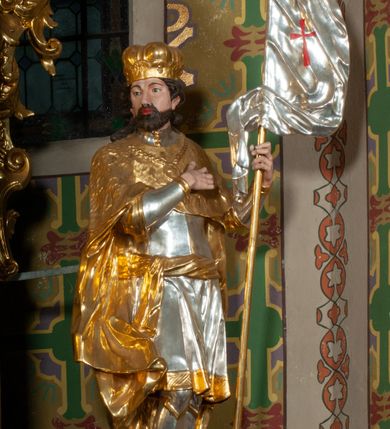 Zdjęcie nr 19: Ołtarz prostopadłościenny z mensą. Antepedium w formie leżącego prostokąta ujętego drewnianą, profilowaną ramą z przedstawieniem Mandylionu i narzędziami Męki Pańskiej na tle wypełnionym ornamentem roślinnym. W centrum, u dołu dwie tarcze herbowe z inicjałami: „TR” i „W”.
Nastawa ołtarzowa architektoniczna, jednoosiowa z bramkami, jednokondygnacyjna ze zwieńczeniem, ustawiona na dwustrefowym cokole. Ujęta dwiema parami kolumn o kapitelach korynckich, ustawionych uskokowo na cokołach zdobionych ornamentem rocaille, dźwigających pełne belkowanie wyłamane na ich osi, nad polem głównym przerwany architraw i fryz oraz gzyms przyjmujący kształt łuku odcinkowego. Na belkowaniu wewnętrznej pary kolumn przerwany wolutowy przyczółek z siedzącymi na nim puttami z narzędziami Męki Pańskiej. W polu głównym wnęka o wykroju prostokąta stojącego zamkniętego łukiem odcinkowym nadwieszonym, w złotej ramie dekorowanej rocaille, w której znajduje się krucyfiks na tle czerwonego atłasu obwieszonego wotami. Powyżej, w partii belkowania rocaillowy kartusz z napisem: „HOC OPUS / NOSTRAE / SALUTIS”. Ołtarz ujęty ażurowymi uszami utworzonymi z ornamentu rocaille.  Zwieńczenie ujęte spływami wolutowymi, zamknięte dwiema skierowanymi do środka wolutami, dekorowane ornamentem rocaille. W polu płycina w formie stojącego owalu wyłożona czerwonym atłasem, ujęta profilowaną ramą z rzeźbą gołębicy Ducha Świętego na tle obłoków w glorii promienistej, flankowana przez akant przechodzący w woluty oraz przez kwiatowe festony z wstęgą. Bramki o wykroju stojącego prostokąta, zamkniętego łukiem odcinkowym nadwieszonym z drzwiczkami, zakończone konsolami ujętymi spływami wolutowymi, stanowiącymi podstawy dla figur św. Michała Archanioła i św. Wacława. Struktura ołtarza malowana na kolor biały, z pozłacaną ornamentyką profilami i kolumnami.

