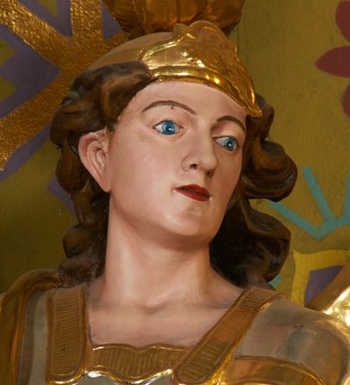 Zdjęcie nr 5: Rzeźba drążona z tyłu, ustawiona na podstawie o nieregularnym planie. Figura ukazana w całej postaci, zwrócona w trzech czwartych w lewą stronę, stoi w kontrapoście. Michał w swojej prawej, uniesionej ręce trzyma miecz skierowany ostrzem w górę, lewą przytrzymuje z boku owalną tarczę z tetragramem („יהוה” - hebrajski czteroliterowy zapis imienia Boga). Ma szeroką twarz o młodzieńczych rysach i migdałowatych oczach, dużym nosie i pełnych ustach, okoloną silnie kręconymi włosami opadającymi na plecy. Ubrany jest w zbroję rzymskiego legionisty z tuniką sięgającą kolan, pancerzem oraz z wysoko zawiązanymi sandałami. Na jego prawym ramieniu zawieszony jest płaszcz opadający na plecy. Na głowie ma hełm z pióropuszem. Skrzydła są złożone po bokach postaci. Polichromia figury naturalistyczna w partiach ciała, tunika i pancerz srebrzone, płaszcz, hełm, sandały, skrzydła, tarcza i detale stroju złocone.

