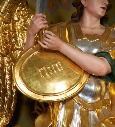 Zdjęcie nr 4: Rzeźba drążona z tyłu, ustawiona na podstawie o nieregularnym planie. Figura ukazana w całej postaci, zwrócona w trzech czwartych w lewą stronę, stoi w kontrapoście. Michał w swojej prawej, uniesionej ręce trzyma miecz skierowany ostrzem w górę, lewą przytrzymuje z boku owalną tarczę z tetragramem („יהוה” - hebrajski czteroliterowy zapis imienia Boga). Ma szeroką twarz o młodzieńczych rysach i migdałowatych oczach, dużym nosie i pełnych ustach, okoloną silnie kręconymi włosami opadającymi na plecy. Ubrany jest w zbroję rzymskiego legionisty z tuniką sięgającą kolan, pancerzem oraz z wysoko zawiązanymi sandałami. Na jego prawym ramieniu zawieszony jest płaszcz opadający na plecy. Na głowie ma hełm z pióropuszem. Skrzydła są złożone po bokach postaci. Polichromia figury naturalistyczna w partiach ciała, tunika i pancerz srebrzone, płaszcz, hełm, sandały, skrzydła, tarcza i detale stroju złocone.

