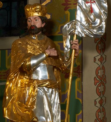 Zdjęcie nr 1: Rzeźba drążona z tyłu, ustawiona na podstawie o nieregularnym planie. Święty ukazany w całej postaci, frontalnie, z głową skierowaną w prawą stronę, stojący w kontrapoście. Prawa ręka postaci spoczywa na piersi, lewą trzyma drzewce zwiniętej chorągwi zakończone krzyżem, na srebrnym płacie chorągwi widoczny czerwony krzyż. Wacław ma szeroką twarz o migdałowatych oczach, dużym nosie i pełnych ustach, okoloną krótką brodą oraz silnie kręconymi włosami opadającymi na ramiona. Ubrany jest w zbroję oraz tunikę widoczną w partii ud, przepasaną w biodrach wstęgą. Ramiona i plecy przykrywa płaszcz z imitującą futro gronostajowe pelerynką, na piersi ma zawieszony łańcuch z krzyżem, na głowie zaś mitrę książęcą. Polichromia figury jest naturalistyczna w partiach ciała, zbroja i tunika srebrzone, płaszcz, mitra i detale stroju złocone. 