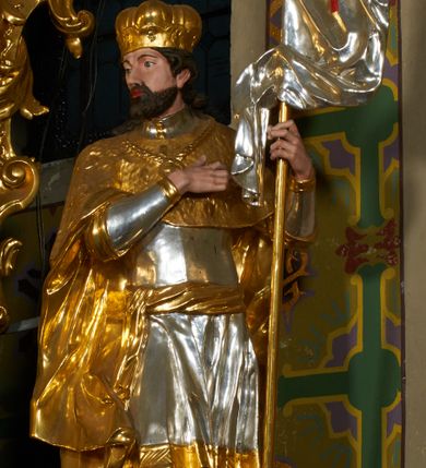 Zdjęcie nr 2: Rzeźba drążona z tyłu, ustawiona na podstawie o nieregularnym planie. Święty ukazany w całej postaci, frontalnie, z głową skierowaną w prawą stronę, stojący w kontrapoście. Prawa ręka postaci spoczywa na piersi, lewą trzyma drzewce zwiniętej chorągwi zakończone krzyżem, na srebrnym płacie chorągwi widoczny czerwony krzyż. Wacław ma szeroką twarz o migdałowatych oczach, dużym nosie i pełnych ustach, okoloną krótką brodą oraz silnie kręconymi włosami opadającymi na ramiona. Ubrany jest w zbroję oraz tunikę widoczną w partii ud, przepasaną w biodrach wstęgą. Ramiona i plecy przykrywa płaszcz z imitującą futro gronostajowe pelerynką, na piersi ma zawieszony łańcuch z krzyżem, na głowie zaś mitrę książęcą. Polichromia figury jest naturalistyczna w partiach ciała, zbroja i tunika srebrzone, płaszcz, mitra i detale stroju złocone. 