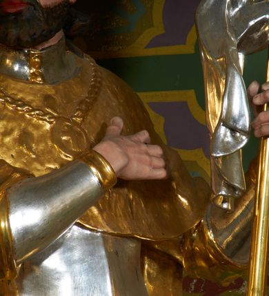 Zdjęcie nr 3: Rzeźba drążona z tyłu, ustawiona na podstawie o nieregularnym planie. Święty ukazany w całej postaci, frontalnie, z głową skierowaną w prawą stronę, stojący w kontrapoście. Prawa ręka postaci spoczywa na piersi, lewą trzyma drzewce zwiniętej chorągwi zakończone krzyżem, na srebrnym płacie chorągwi widoczny czerwony krzyż. Wacław ma szeroką twarz o migdałowatych oczach, dużym nosie i pełnych ustach, okoloną krótką brodą oraz silnie kręconymi włosami opadającymi na ramiona. Ubrany jest w zbroję oraz tunikę widoczną w partii ud, przepasaną w biodrach wstęgą. Ramiona i plecy przykrywa płaszcz z imitującą futro gronostajowe pelerynką, na piersi ma zawieszony łańcuch z krzyżem, na głowie zaś mitrę książęcą. Polichromia figury jest naturalistyczna w partiach ciała, zbroja i tunika srebrzone, płaszcz, mitra i detale stroju złocone. 