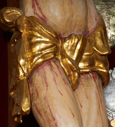 Zdjęcie nr 2: Krzyż drewniany o prostych ramionach z figurą w typie Cristo vivo przybitą trzema gwoździami. Sylwetka w lekkim zwisie, o szerokim rozłożeniu ramion, z głową przechyloną na prawe ramię; nogi ugięte w kolanach, skierowane w prawą stronę, stopy w układzie prawa na lewą. Twarz pociągła o wąskich, otwartych oczach, dużym nosie, otwartych ustach, okolona brodą oraz włosami skręconymi w rurkowate pukle. Na głowie korona cierniowa, wokół niej trzy wiązki promieni. Ciało wychudzone, o zaznaczonych mięśniach ramion i nóg oraz sumarycznie kształtowanej klatce piersiowej z podkreślonymi liniami żeber i wypukłością brzucha oraz ze śladami męki w postaci rzeźbionych kropel krwi wypływającej z ran. Perizonium krótkie, ściśle opinające biodra, zawiązane w partii podbrzusza, ze zwisem na prawym biodrze. Na górnym zakończeniu pionowej belki krzyża banderola z napisem „INRI”. Ciało polichromowane naturalistycznie, perizonium pozłacane. 
