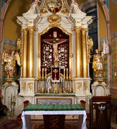 Zdjęcie nr 5: Krzyż drewniany o prostych ramionach z figurą w typie Cristo vivo przybitą trzema gwoździami. Sylwetka w lekkim zwisie, o szerokim rozłożeniu ramion, z głową przechyloną na prawe ramię; nogi ugięte w kolanach, skierowane w prawą stronę, stopy w układzie prawa na lewą. Twarz pociągła o wąskich, otwartych oczach, dużym nosie, otwartych ustach, okolona brodą oraz włosami skręconymi w rurkowate pukle. Na głowie korona cierniowa, wokół niej trzy wiązki promieni. Ciało wychudzone, o zaznaczonych mięśniach ramion i nóg oraz sumarycznie kształtowanej klatce piersiowej z podkreślonymi liniami żeber i wypukłością brzucha oraz ze śladami męki w postaci rzeźbionych kropel krwi wypływającej z ran. Perizonium krótkie, ściśle opinające biodra, zawiązane w partii podbrzusza, ze zwisem na prawym biodrze. Na górnym zakończeniu pionowej belki krzyża banderola z napisem „INRI”. Ciało polichromowane naturalistycznie, perizonium pozłacane. 
