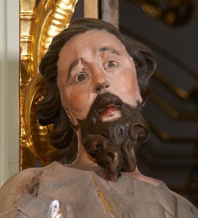 Zdjęcie nr 7: Rzeźba z tyłu drążona, ustawiona na niewielkiej, prostej podstawie. Joachim przedstawiony w całej postaci, ustawiony  frontalnie, z głową skierowaną w prawo, stojąc w kontrapoście, odchylony jest delikatnie w tył. Prawa dłoń świętego spoczywa na piersi, lewa wyciągnięta jest do przodu, w geście przemowy. Twarz świętego jest szczupła i pociągła, o dużych oczach z głęboko rzeźbionymi powiekami oraz uniesionymi brwiami. Nos ma prosty, usta pełne, rozchylone, okolona jest gęstą, kręconą brodą oraz włosami sięgającymi ramion, układającymi się w głęboko rzeźbione pukle. Święty ubrany jest w długą tunikę przepasaną w talii, obficie drapowaną i przylegającą do ciała oraz w płaszcz opasujący diagonalnie postać, zawieszony na lewym przedramieniu świętego z rozwianym zwisem tkaniny na lewym boku. Figura polichromowana naturalistycznie w partiach ciała, broda i włosy brązowe, tunika srebrzona, płaszcz złocony.
