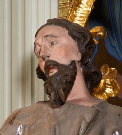 Zdjęcie nr 5: Rzeźba z tyłu drążona, ustawiona na niewielkiej, prostej podstawie. Joachim przedstawiony w całej postaci, ustawiony  frontalnie, z głową skierowaną w prawo, stojąc w kontrapoście, odchylony jest delikatnie w tył. Prawa dłoń świętego spoczywa na piersi, lewa wyciągnięta jest do przodu, w geście przemowy. Twarz świętego jest szczupła i pociągła, o dużych oczach z głęboko rzeźbionymi powiekami oraz uniesionymi brwiami. Nos ma prosty, usta pełne, rozchylone, okolona jest gęstą, kręconą brodą oraz włosami sięgającymi ramion, układającymi się w głęboko rzeźbione pukle. Święty ubrany jest w długą tunikę przepasaną w talii, obficie drapowaną i przylegającą do ciała oraz w płaszcz opasujący diagonalnie postać, zawieszony na lewym przedramieniu świętego z rozwianym zwisem tkaniny na lewym boku. Figura polichromowana naturalistycznie w partiach ciała, broda i włosy brązowe, tunika srebrzona, płaszcz złocony.
