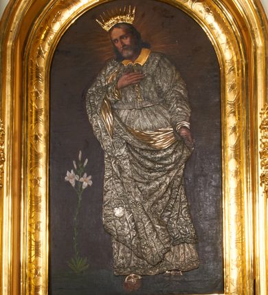 Zdjęcie nr 1: Obraz w kształcie stojącego prostokąta, zamkniętego łukiem półkolistym. W centrum kompozycji ukazany św. Józef w postawie stojącej, delikatnie zwrócony w prawą stronę, z prawą stopą wysuniętą do przodu a prawą ręką złożoną na piersi, zaś lewą ręką przytrzymujący połę płaszcza u boku. Twarz świętego owalna, z długim i prostym nosem, okolona siwą brodą oraz długimi i delikatnie siwymi włosami, opadającymi na ramiona. Postać przykryta metalową sukienką w kolorze srebra, na powierzchni której oddano przebieg draperii szat świętego oraz zdobiący je ornament roślinny, a nad głową złoconą, kameryzowaną i promienistą koronę. Tło jednolite, brunatne, wokół głowy św. Józefa delikatnie rozjaśnione na kształt nimbu. Po lewej stronie obrazu krzak białej lilii. 


