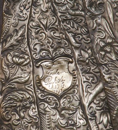 Zdjęcie nr 5: Obraz w kształcie stojącego prostokąta, zamkniętego łukiem półkolistym. W centrum kompozycji ukazany św. Józef w postawie stojącej, delikatnie zwrócony w prawą stronę, z prawą stopą wysuniętą do przodu a prawą ręką złożoną na piersi, zaś lewą ręką przytrzymujący połę płaszcza u boku. Twarz świętego owalna, z długim i prostym nosem, okolona siwą brodą oraz długimi i delikatnie siwymi włosami, opadającymi na ramiona. Postać przykryta metalową sukienką w kolorze srebra, na powierzchni której oddano przebieg draperii szat świętego oraz zdobiący je ornament roślinny, a nad głową złoconą, kameryzowaną i promienistą koronę. Tło jednolite, brunatne, wokół głowy św. Józefa delikatnie rozjaśnione na kształt nimbu. Po lewej stronie obrazu krzak białej lilii. 


