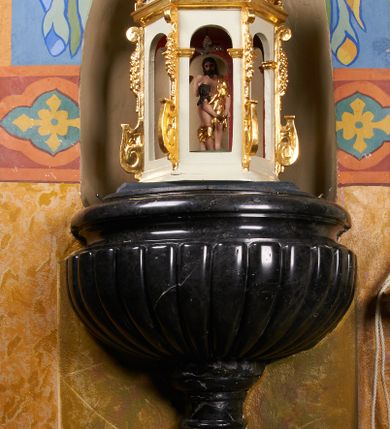 Zdjęcie nr 1: Chrzcielnica osadzona na wysokim postumencie wmurowanym w ścianę, prostopadłościennym cokole, z trzonem w typie gruszkowym oraz czarą z puklami i profilowanym brzegiem. Na trzonie herb Przyjaciel i sygle wokół niego: &quot;MB O[LSZYŃSKI] / S[ANCTAE] T[HEOLOGIAE] D[OCTOR] / P[AROCHUS] I[GOLOMIENSIS]&quot;. Pokrywa w kształcie ażurowej świątynki, na rzucie sześcioboku, ujętej spływami wolutowymi, nakryta czaszą ozdobioną liśćmi akantu, z popiersiem Boga Ojca błogosławiącego z kulą w dłoni; wewnątrz św. Jana Chrzciciel chrzczący Jezusa w Jordanie.  