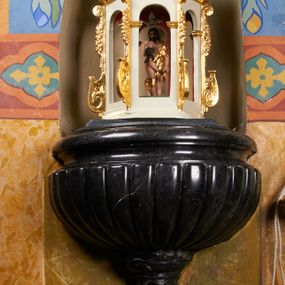 Zdjęcie nr 1: Chrzcielnica osadzona na wysokim postumencie wmurowanym w ścianę, prostopadłościennym cokole, z trzonem w typie gruszkowym oraz czarą z puklami i profilowanym brzegiem. Na trzonie herb Przyjaciel i sygle wokół niego: "MB O[LSZYŃSKI] / S[ANCTAE] T[HEOLOGIAE] D[OCTOR] / P[AROCHUS] I[GOLOMIENSIS]". Pokrywa w kształcie ażurowej świątynki, na rzucie sześcioboku, ujętej spływami wolutowymi, nakryta czaszą ozdobioną liśćmi akantu, z popiersiem Boga Ojca błogosławiącego z kulą w dłoni; wewnątrz św. Jana Chrzciciel chrzczący Jezusa w Jordanie.  