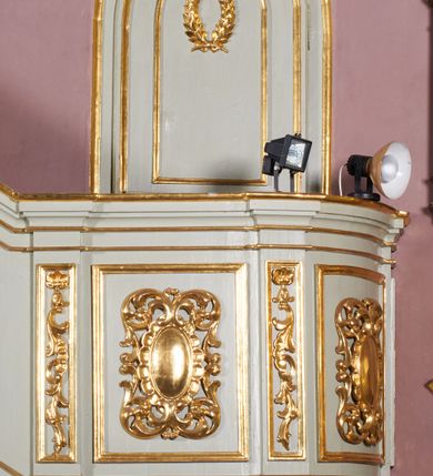 Zdjęcie nr 2: Ambona wisząca, przyścienna, z dostępem od strony północnej w formie podestu z balustradą, prowadzącego do drzwi przekutych w murze. Kosz na planie zbliżonym do kwadratu, o wklęsło-wypukłych ściankach, na cokole dekorowanym rocaille'em, z podwieszeniem w formie stożka ujętego spływami wolutowymi i zakończonego szyszką. Ścianki kosza ujęte w narożach pilastrami o trzonach w formie spływów wolutowych dekorowanych kampanulami, podtrzymujących belkowanie z gzymsem tworzącym parapet. Na ściankach płyciny zdobione rocaille'em. Balustrada podestu pełna, dekorowana płycinami ze stylizowanym ornamentem roślinnym. Zaplecek flankowany pilastrami o kanelowanych trzonach, o kapitelach w formie konsol dźwigających imposty oraz fragmenty przerwanego, wolutowego, odwróconego na zewnątrz przyczółka. W polu zaplecka płycina w kształcie stojącego prostokąta o wklęsłych narożach, z malowanym krzyżem ujętym wieńcem laurowym i tablicami Dekalogu. Zaplecek flankowany uszami w formie stylizowanego rocaille'u. Całość zwieńczona rzeźbą gołębicy Ducha Świętego w glorii promienistej. Struktura ambony polichromowana na kolor biały, profile i ornamentyka złocone, detale srebrzone. 