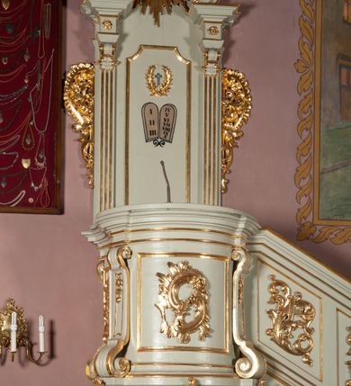 Zdjęcie nr 6: Ambona wisząca, przyścienna, z dostępem od strony północnej w formie podestu z balustradą, prowadzącego do drzwi przekutych w murze. Kosz na planie zbliżonym do kwadratu, o wklęsło-wypukłych ściankach, na cokole dekorowanym rocaille'em, z podwieszeniem w formie stożka ujętego spływami wolutowymi i zakończonego szyszką. Ścianki kosza ujęte w narożach pilastrami o trzonach w formie spływów wolutowych dekorowanych kampanulami, podtrzymujących belkowanie z gzymsem tworzącym parapet. Na ściankach płyciny zdobione rocaille'em. Balustrada podestu pełna, dekorowana płycinami ze stylizowanym ornamentem roślinnym. Zaplecek flankowany pilastrami o kanelowanych trzonach, o kapitelach w formie konsol dźwigających imposty oraz fragmenty przerwanego, wolutowego, odwróconego na zewnątrz przyczółka. W polu zaplecka płycina w kształcie stojącego prostokąta o wklęsłych narożach, z malowanym krzyżem ujętym wieńcem laurowym i tablicami Dekalogu. Zaplecek flankowany uszami w formie stylizowanego rocaille'u. Całość zwieńczona rzeźbą gołębicy Ducha Świętego w glorii promienistej. Struktura ambony polichromowana na kolor biały, profile i ornamentyka złocone, detale srebrzone. 