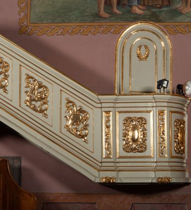 Zdjęcie nr 9: Ambona wisząca, przyścienna, z dostępem od strony północnej w formie podestu z balustradą, prowadzącego do drzwi przekutych w murze. Kosz na planie zbliżonym do kwadratu, o wklęsło-wypukłych ściankach, na cokole dekorowanym rocaille'em, z podwieszeniem w formie stożka ujętego spływami wolutowymi i zakończonego szyszką. Ścianki kosza ujęte w narożach pilastrami o trzonach w formie spływów wolutowych dekorowanych kampanulami, podtrzymujących belkowanie z gzymsem tworzącym parapet. Na ściankach płyciny zdobione rocaille'em. Balustrada podestu pełna, dekorowana płycinami ze stylizowanym ornamentem roślinnym. Zaplecek flankowany pilastrami o kanelowanych trzonach, o kapitelach w formie konsol dźwigających imposty oraz fragmenty przerwanego, wolutowego, odwróconego na zewnątrz przyczółka. W polu zaplecka płycina w kształcie stojącego prostokąta o wklęsłych narożach, z malowanym krzyżem ujętym wieńcem laurowym i tablicami Dekalogu. Zaplecek flankowany uszami w formie stylizowanego rocaille'u. Całość zwieńczona rzeźbą gołębicy Ducha Świętego w glorii promienistej. Struktura ambony polichromowana na kolor biały, profile i ornamentyka złocone, detale srebrzone. 