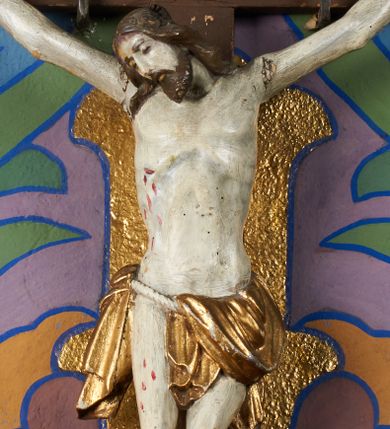 Zdjęcie nr 1: Do krzyża o prostych ramionach, zwieńczonego tabliczką z napisem „I.N./R.I.”, przybita jest trzema gwoździami figura umarłego Chrystusa. Jego ciało zostało ukazane w średnim zwisie, o nieznacznie łukowatym układzie ramion, głowa opada na prawy bark, lewa noga jest ugięta w kolanie, prawa wyprostowana, stopy w układzie prawa na lewą. Chrystus ma pociągłą twarz, o zamkniętych oczach, okoloną krótką brodą i brązowymi, falowanymi włosami opadającymi na ramiona. Jego ciało jest stosunkowo szczupłe, o szerokiej, umięśnionej klatce piersiowej z widocznym zarysem żeber oraz podkreślonej muskulaturze i ścięgnach ramion. Perizonium zawieszone na sznurze, odsłania prawe udo. Polichromia karnacji naturalistyczna z zaznaczonymi śladami męki, perizonium pozłacane. 