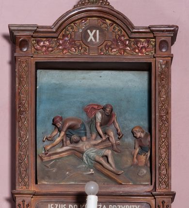 Zdjęcie nr 3: Stacje Drogi krzyżowej w kształcie stojących prostokątów zostały ujęte architektoniczną ramą. Obrazy ustawiono na cokole, po bokach są flankowane dwoma pilastrami, podtrzymującymi wyłamane belkowanie, zamknięte gzymsem w kształcie łuku nadwieszonego. Na szczycie, w rzeźbionych owocach winogron osadzono grecki krzyżyk o trójlistnie zakończonych ramionach z uskokami. Pilastry są zdobione symetryczną wicią cierniową, a pole zwieńczenia bogato dekorowane wicią roślinną złożoną z bujnych, czerwonych kwiatów otaczających numer stacji umieszczony w owalu na tle krzyżyka greckiego. 
Sceny przeważnie wielopostaciowe, płaskorzeźbione, barwnie polichromowane. Postacie o poprawnej anatomii ciała, poruszone, na pierwszym planie bardziej wypukłe. Tło błękitne, malowane. W partii cokołu umieszczono malowane tytuły stacji:
I. „JEZUS NA ŚMIERĆ OSĄDZONY.”
II. „JEZUS BIERZE KRZYŻ NA RAMIONA.”
III. „JEZUS PIERWSZY RAZ UPADA.”
IV. „JEZUS SPOTYKA NAJŚW(IĘTSZĄ) MATKĘ SWOJĄ”
V. „SZYMON CYR(ENEJCZYK) POMAGA NIEŚĆ KRZYŻ JEZUSA”
VI. „ŚW(IĘTA) WERONIKA OCIERA TWARZ JEZUSA”
VII. „JEZUS DRUGI RAZ UPADA”
VIII. „JEZUS NAPOTYKA PŁACZĄCE NIEWIASTY”
IX. „JEZUS TRZECI RAZ UPADA.”
X. „JEZUS Z SZAT OBNAŻONY”
XI. „JEZUS DO KRZYŻA PRZYBITY”
XII. „JEZUS NA KRZYŻU UMIERA.”
XIII. „JEZUS Z KRZYŻA ZDJĘTY.”
XIV. „JEZUS DO GROBU ZŁOŻONY.”
