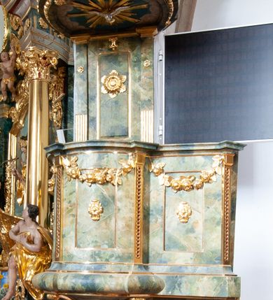 Zdjęcie nr 3: Ambona przyścienna, wisząca, z dostępem od strony prezbiterium w formie podestu i jednobiegowych schodów z pełną balustradą. Kosz na rzucie ściętego koła, na spłaszczonym wałku, z podwieszeniem o formie zbliżonej do stożka o wklęsłych bokach, ujętego czterema wolutami i pierścieniem, zakończonego szyszką. Ścianki kosza wydzielone wąskimi pilastrami, o trzonach dekorowanych plecionką, dźwigającymi gzyms stanowiący parapet, wyłamany na ich osi. W ich polach płyciny z rozetami, pod gzymsem podwieszone kwiatowe girlandy. Zaplecek wąski, flankowany dwoma pilastrami, kanelowanymi w dolnej partii trzonów, z rozetami w górnej. W polu płycina w formie stojącego prostokąta z dużą rozetą w środku, nad nią klucz. Baldachim na planie analogicznym do kosza, dekorowany kimationem oraz ornamentem roślinnym. Na nim rzeźbiony fragment skały z dwiema uskrzydlonymi główkami anielskimi, na którym leży otwarta księga z napisem „POENITE / MINI ET / CREDITE / EVANGE/LIO Marci 1. v(ersum) 15.”; za nią ustawione liście palm i duży krzyż, z promieniami na skrzyżowaniu belek. Obok, na skale siedzi putto, za nim gorejące serce i kotwica. Na podniebieniu baldachimu gołębica Ducha Świętego w glorii promienistej. Struktura ambony polichromowana na sposób imitujący marmoryzację w kolorze żółto-zielonym, detale i ornamentyka złocone. 
