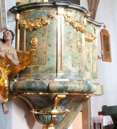 Zdjęcie nr 1: Ambona przyścienna, wisząca, z dostępem od strony prezbiterium w formie podestu i jednobiegowych schodów z pełną balustradą. Kosz na rzucie ściętego koła, na spłaszczonym wałku, z podwieszeniem o formie zbliżonej do stożka o wklęsłych bokach, ujętego czterema wolutami i pierścieniem, zakończonego szyszką. Ścianki kosza wydzielone wąskimi pilastrami, o trzonach dekorowanych plecionką, dźwigającymi gzyms stanowiący parapet, wyłamany na ich osi. W ich polach płyciny z rozetami, pod gzymsem podwieszone kwiatowe girlandy. Zaplecek wąski, flankowany dwoma pilastrami, kanelowanymi w dolnej partii trzonów, z rozetami w górnej. W polu płycina w formie stojącego prostokąta z dużą rozetą w środku, nad nią klucz. Baldachim na planie analogicznym do kosza, dekorowany kimationem oraz ornamentem roślinnym. Na nim rzeźbiony fragment skały z dwiema uskrzydlonymi główkami anielskimi, na którym leży otwarta księga z napisem „POENITE / MINI ET / CREDITE / EVANGE/LIO Marci 1. v(ersum) 15.”; za nią ustawione liście palm i duży krzyż, z promieniami na skrzyżowaniu belek. Obok, na skale siedzi putto, za nim gorejące serce i kotwica. Na podniebieniu baldachimu gołębica Ducha Świętego w glorii promienistej. Struktura ambony polichromowana na sposób imitujący marmoryzację w kolorze żółto-zielonym, detale i ornamentyka złocone. 
