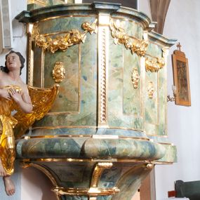 Zdjęcie nr 1: Ambona przyścienna, wisząca, z dostępem od strony prezbiterium w formie podestu i jednobiegowych schodów z pełną balustradą. Kosz na rzucie ściętego koła, na spłaszczonym wałku, z podwieszeniem o formie zbliżonej do stożka o wklęsłych bokach, ujętego czterema wolutami i pierścieniem, zakończonego szyszką. Ścianki kosza wydzielone wąskimi pilastrami, o trzonach dekorowanych plecionką, dźwigającymi gzyms stanowiący parapet, wyłamany na ich osi. W ich polach płyciny z rozetami, pod gzymsem podwieszone kwiatowe girlandy. Zaplecek wąski, flankowany dwoma pilastrami, kanelowanymi w dolnej partii trzonów, z rozetami w górnej. W polu płycina w formie stojącego prostokąta z dużą rozetą w środku, nad nią klucz. Baldachim na planie analogicznym do kosza, dekorowany kimationem oraz ornamentem roślinnym. Na nim rzeźbiony fragment skały z dwiema uskrzydlonymi główkami anielskimi, na którym leży otwarta księga z napisem „POENITE / MINI ET / CREDITE / EVANGE/LIO Marci 1. v(ersum) 15.”; za nią ustawione liście palm i duży krzyż, z promieniami na skrzyżowaniu belek. Obok, na skale siedzi putto, za nim gorejące serce i kotwica. Na podniebieniu baldachimu gołębica Ducha Świętego w glorii promienistej. Struktura ambony polichromowana na sposób imitujący marmoryzację w kolorze żółto-zielonym, detale i ornamentyka złocone. 
